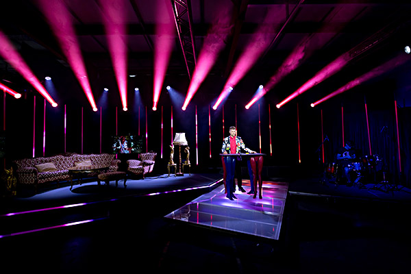 Stage with pink-purple lighting, a presenter stands at a high table, sofas and lamps as stage decoration in the background.
