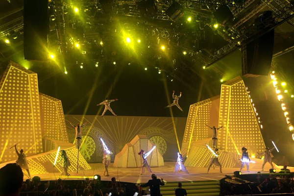 Große Showbühne in Saudi-Arabien mit gelben Lichteffekten. Akrobaten hängen in der Luft und Performer mit leuchtenden Stäben stehen auf der Bühne, umgeben von futuristischen Kulissen.