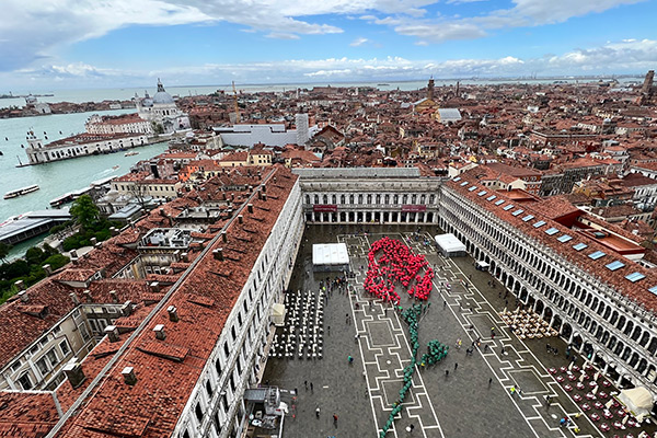 Luftaufnahme des Markusplatzes in Venedig mit einer großen Kunstinstallation in Form einer roten Rose, die aus Menschen gebildet wird.