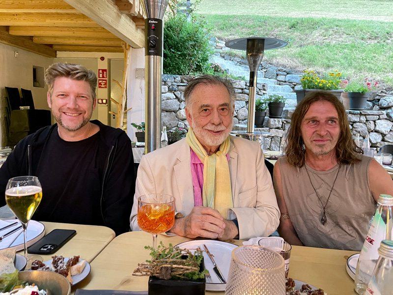 Drei Männer sitzen an einem Esstisch im Freien, lächeln in die Kamera. Vor ihnen stehen Getränke und Teller mit Speisen. Im Hintergrund eine Steinmauer und Grünfläche.