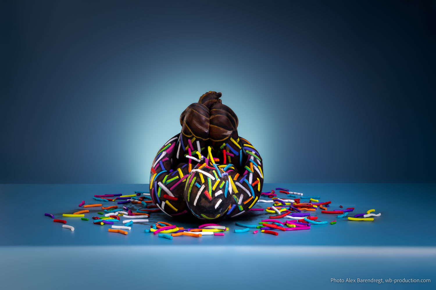 Ein in Schokoladenoptik bemalter Körper ist mit bunten Zuckerstreuseln dekoriert und sitzt vor blauem Hintergrund.