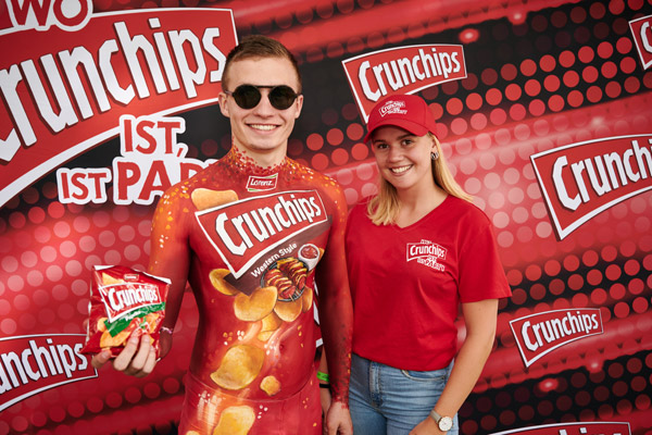 Mann mit Crunchips-Bodypainting und Chipstüte posiert mit einer Frau im roten Crunchips-T-Shirt vor einer gebrandeten Fotowand.