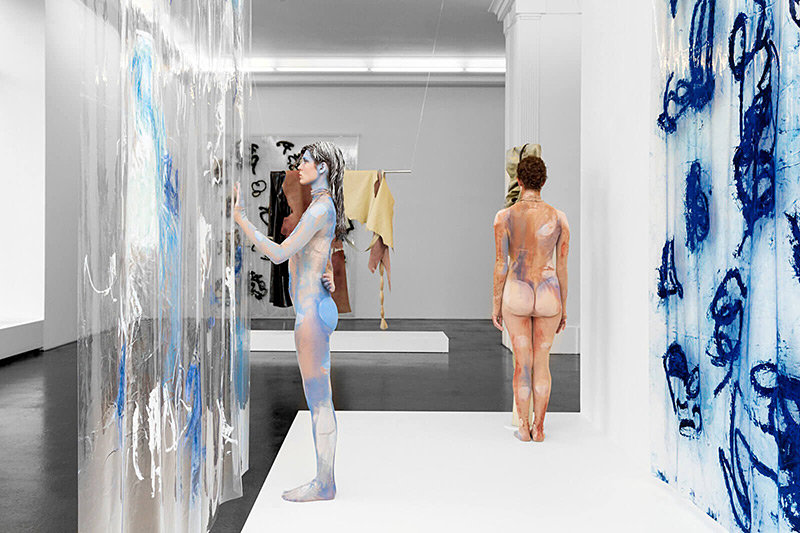 Zwei nackte Frauen mit blauer und beiger Körperbemalung stehen in einer modernen Galerie. Eine betrachtet eine transparente Wand mit Malereien, während die andere mit dem Rücken zur Kamera steht.