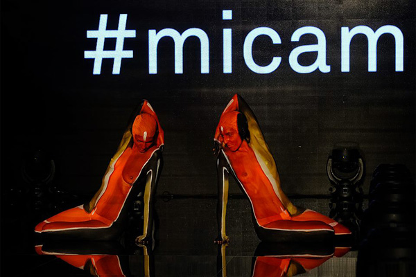 Zwei Models sind als High Heels bemalt und posieren auf einer Bühne vor einer schwarzen Wand mit dem weißen Schriftzug „#micam“. Die Körperbemalung lässt sie wie große, rot-schwarze Schuhe aussehen.