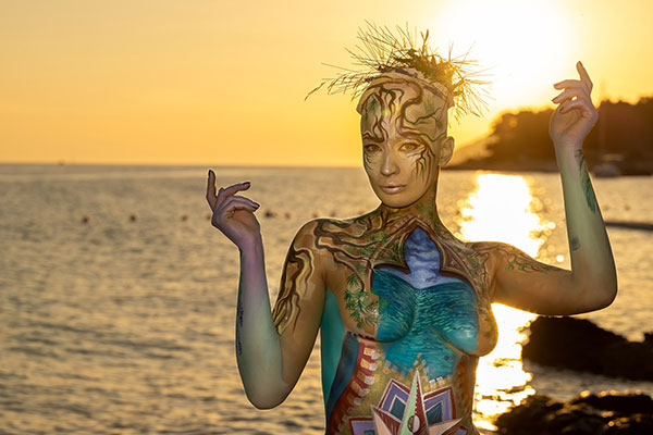 Körperkunstmodell in fantasievollem Bodypainting vor Sonnenuntergang am Meer, mit goldenem Licht im Hintergrund.