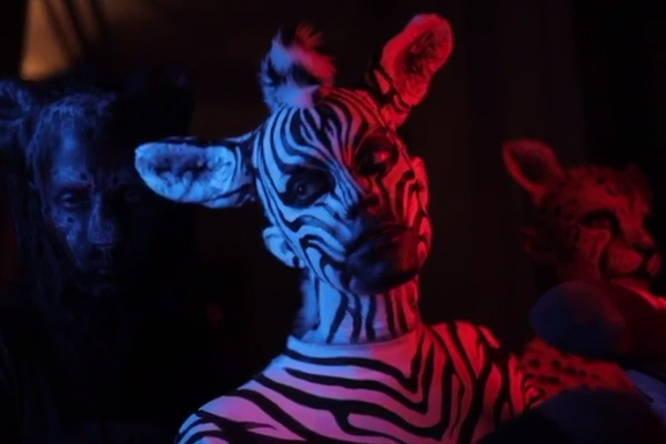 Person mit Bodypainting im Zebramuster, beleuchtet in rotem und blauem Licht, flankiert von weiteren Tierdarstellungen im Hintergrund.