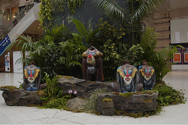Bodypainting-Installation in einem Einkaufszentrum mit Menschen, die realistische Mandrill-Affen darstellen und in einer tropischen Kulisse sitzen.