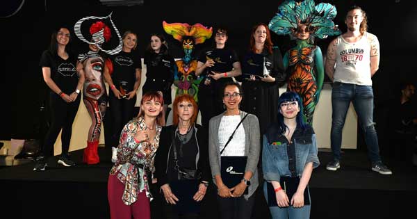 Gruppenfoto bei einem Bodypainting-Wettbewerb: Künstler und Models mit fantasievollen, farbenprächtigen Bodypaintings posieren gemeinsam auf der Bühne, während Jury und Gäste davor stehen.