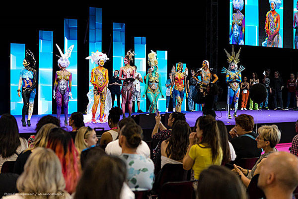Models in farbenfrohem Bodypainting und aufwendigen Kopfschmuck stehen nebeneinander auf einer Bühne bei einer Bodypainting-Show, während das Publikum aufmerksam zusieht und fotografiert.