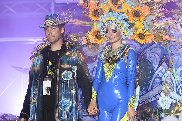 Ein Mann hält auf einer Bühne die Hand eines Models, das in einem blau-gelben Bodypainting mit aufwendigem Kopfschmuck aus Sonnenblumen und Blumen geschmückt ist. Beide präsentieren sich beim Bodypainting-Festival.