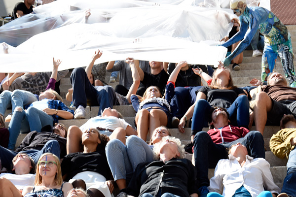 Eine Gruppe von Menschen liegt auf einer Treppe und hält ein großes transparentes Tuch über sich. Rechts steht eine bemalte Person im Bodypainting-Kostüm und hilft beim Ausbreiten des Stoffes. Die Szene wirkt wie eine Performance oder Kunstaktion im Freien.