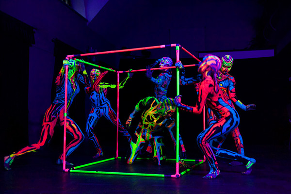 Tänzer in einem Neon-Bodypainting unter UV-Licht, die in und um einen leuchtenden Würfelrahmen performen.
