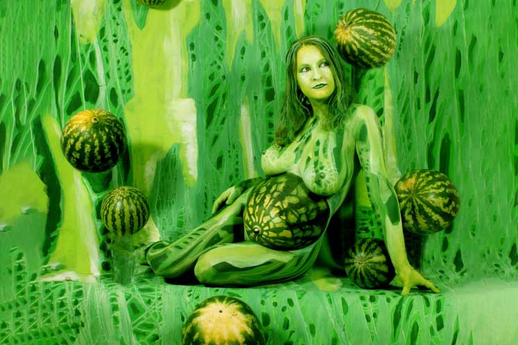 In einer verschneiten Winterlandschaft steht ein bemalter Körper, der sich perfekt in die Form und Struktur eines Baumes einfügt, mit erhobenen Armen wie verschmolzen mit den schneebedeckten Ästen.