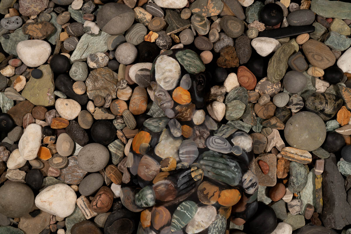 Ein kunstvoll bemalter Körper verschmilzt mit einem Hintergrund aus verschiedenfarbigen Steinen und wirkt fast unsichtbar in der steinigen Umgebung.