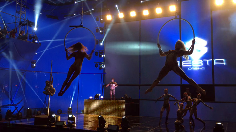 Show im Casino: Akrobaten führen eine Luftakrobatik an Ringen auf, während weitere Tänzer auf der Bühne auftreten. Im Hintergrund erleuchtet eine große LED-Wand mit Casino-Logo die Szene.
