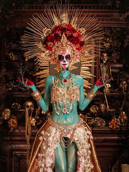 Frau mit türkis bemaltem Körper, aufwendigem Totenkopf-Makeup und goldener Krone mit roten Rosen in prächtigem Outfit.