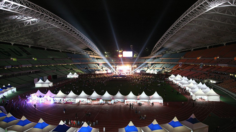 Großes Nachtevent in einem Stadion: Zahlreiche weiße Zelte säumen die Laufbahn, während auf der hell erleuchteten Bühne im Zentrum eine Show stattfindet. Besucher füllen die Arena.