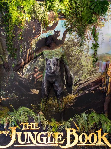 Bodypainting-Illusion zu Disneys ‚The Jungle Book‘: Eine Person ist so bemalt, dass sie fast unsichtbar in einem Dschungel-Backdrop verschmilzt. Auf dem Körper ist der Bär Balu dargestellt, während um sie herum die Filmfiguren und üppige Vegetation zu sehen sind.