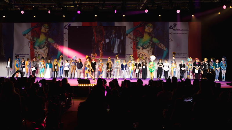 Bühnenauftritt bei einer Bodypainting-Veranstaltung: Eine große Gruppe von Künstlern und Models steht in einer Reihe auf der beleuchteten Bühne, während das Publikum von den Rängen zusieht.