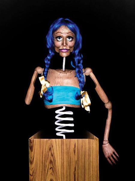 Künstlerische Bodypainting-Illusion: Eine Person ist so bemalt, dass sie wie eine Holzpuppe mit blauen Zöpfen aussieht, die als Jack-in-the-Box-Figur aus einer hölzernen Spielzeugkiste mit Spiralfeder herausragt.