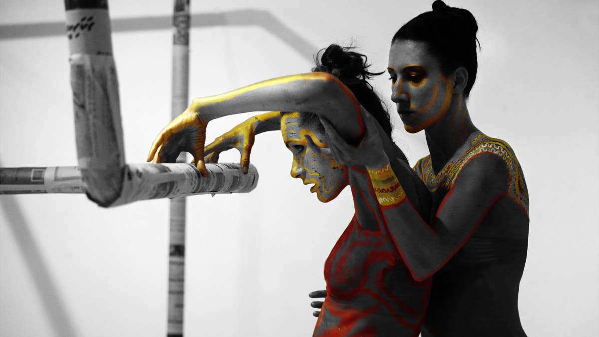 Künstlerische Szene in einer Galerie: Zwei Frauen mit rot-goldenem Bodypainting stehen eng beieinander. Eine Figur lehnt sich nach vorn und hält sich an einer Stange fest, die mit Zeitungspapier umwickelt ist, während die andere sie stützt