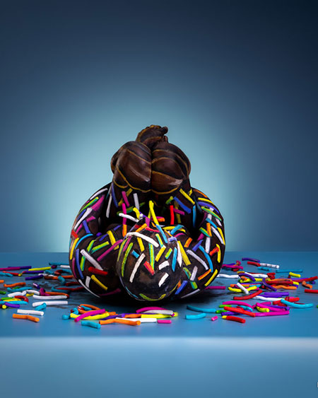 Ein in Schokoladenoptik bemalter Körper ist mit bunten Zuckerstreuseln dekoriert und sitzt vor blauem Hintergrund.