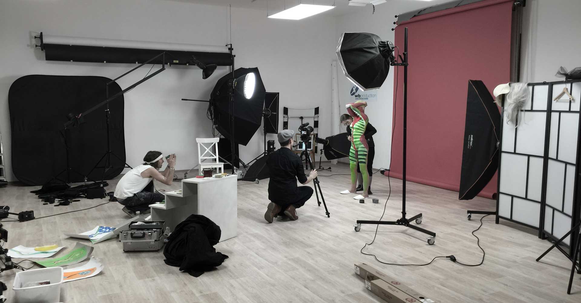 Making-of-Foto im Fotostudio: Ein Kamerateam filmt ein Bodypainting-Shooting, bei dem ein Model in grünen Streifen vor einem roten Hintergrund posiert.