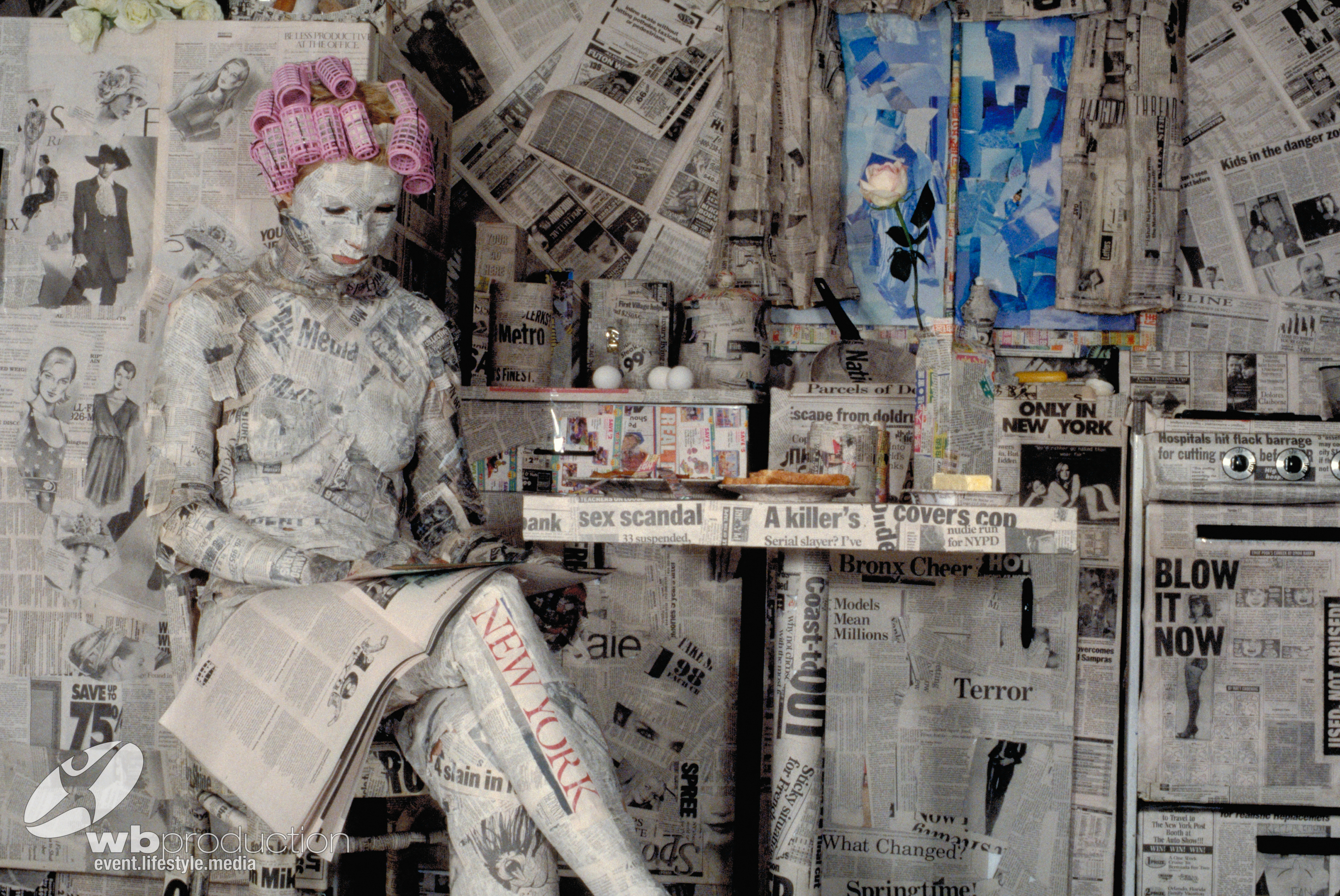 Bodypainting-Kunst: Eine Person, vollständig in Zeitungsdruck bemalt, sitzt in einer mit Zeitungen gestalteten Küche und liest eine Zeitung.