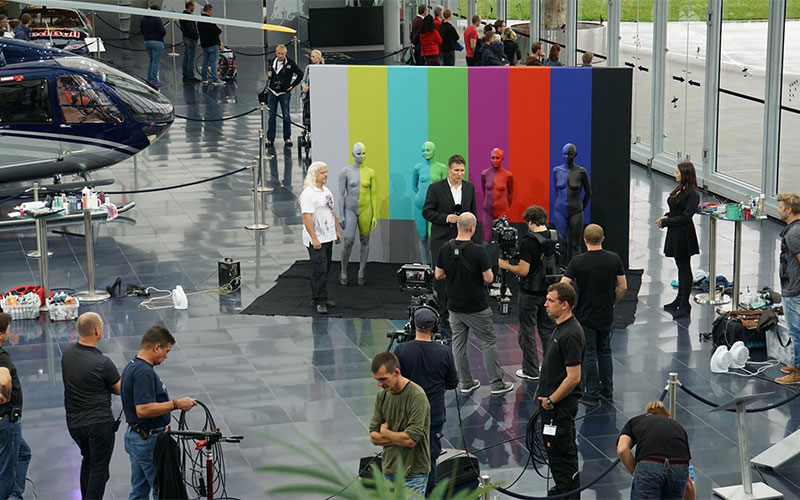 Making-of-Szene in einer Ausstellungshalle: Vor einer großen Testbild-Wand stehen mehrere Models, die jeweils in unterschiedlichen Farben bemalt sind. Kamerateams filmen die Szene, während Zuschauer und Crew im Hintergrund zu sehen sind.